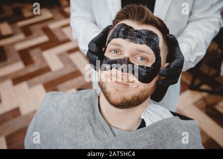 Spa e cura della pelle per gli uomini. Barber Professional applica maschera di carbone nero all'uomo del cliente Foto Stock