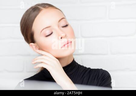 Ritratto bella ragazza con trucco e estensioni ciglia sul labbro rosa, giacca nera su sfondo bianco mattone. Concetto di cura della pelle Foto Stock