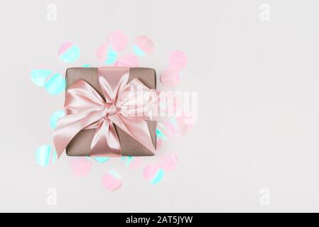 Confezione regalo confezionata in carta artigianale con nastro rosa perfetto su sfondo rustico rosa pallido con confetti olografici. Foto Stock