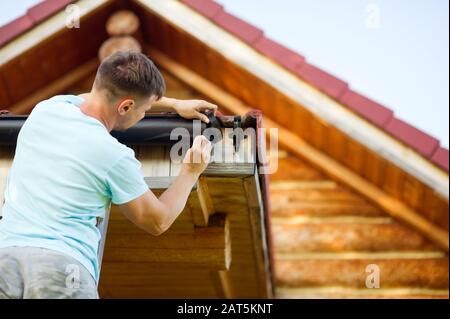 Un uomo in una maglietta bianca fissa elementi sul tetto di una casa di legno Foto Stock