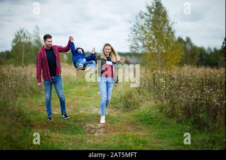 i genitori nei vestiti casuali camminano con il loro piccolo figlio lungo una strada di campagna, tenendo le mani e aiutandolo a rimbalzare Foto Stock