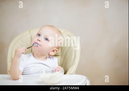 la bambina mangia il porridge di riso con un cucchiaio di metallo e guarda sognatamente davanti a se stessa, copia spazio Foto Stock