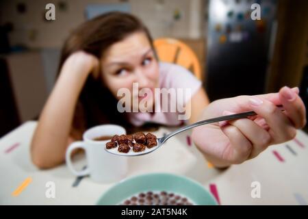 Una giovane donna mostra un cucchiaio con palline di cioccolato ed esprime una riluttanza a mangiarle. Cereali al cioccolato con latte a fuoco e ragazza con grimace in Foto Stock