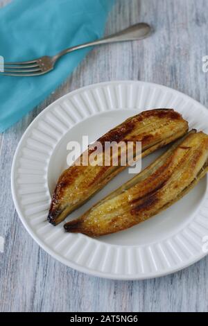 La banana tagliata a metà e cotta nella sua pelle fino al marrone dorato, servita su un piatto bianco. Scena con forchetta e tovagliolo blu Foto Stock