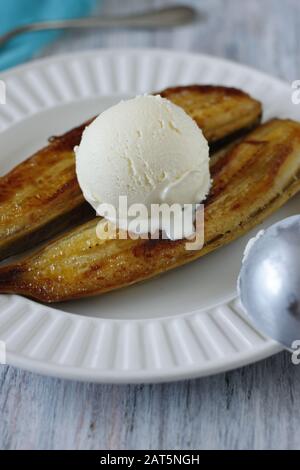 Banana cotta al forno dorato tagliata a metà con copertura di pelle con una paletta di gelato alla vaniglia su piastra bianca. Foto Stock