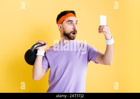 sportivo scioccato lavorando con un manubrio pesante e prendendo selfie isolato su giallo Foto Stock