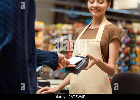 Shopping Giornaliero. Uomo in piedi al banco del supermercato pagare per gli acquisti con smartphone Contactless primo piano cassiere sorridente allegro Foto Stock
