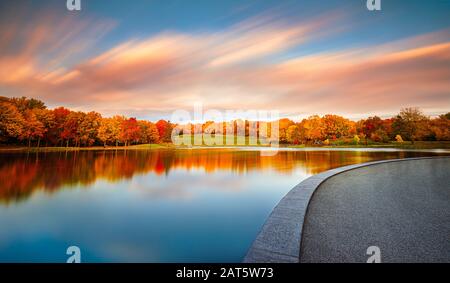 Il bel fogliame arancione dell'autunno e il cielo blu con striature e nuvole colorate si riflettono nella superficie perfettamente ferma del lago. Foto Stock