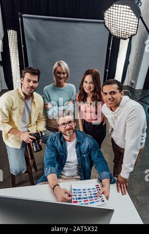 vista dall'alto di un allegro direttore creativo e di assistenti che guardano la fotocamera in uno studio fotografico Foto Stock