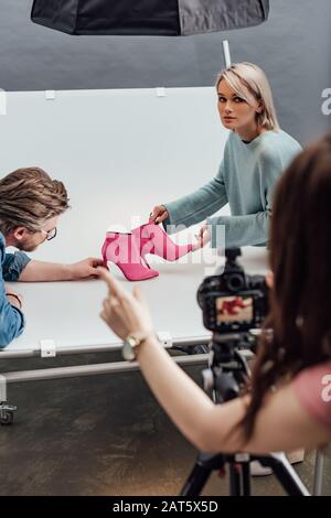 vista dal taglio corto del fotografo che punta con un dito vicino a un assistente attraente e a un direttore artistico che tiene scarpe rosa Foto Stock