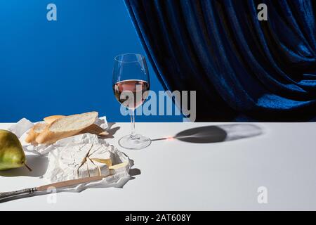 Classica ancora vita con pera, vino rosso, baguette e formaggio Camembert su tavola bianca vicino velour tenda isolato su blu Foto Stock