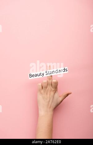 vista corta della donna che mostra il dito medio e la carta con gli standard di bellezza scritta su sfondo rosa Foto Stock