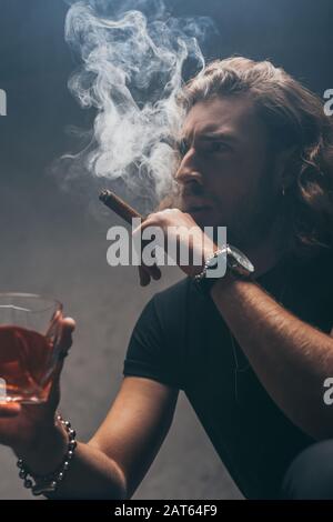 uomo d'affari alla moda teso in abito nero fumo sigaro e bere whisky vicino alla parete grigia Foto Stock