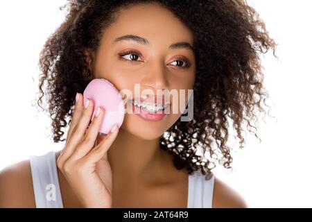 attraente ragazza africana americana felice con bretelle usando la spazzola facciale di pulizia del silicone, isolata su bianco Foto Stock