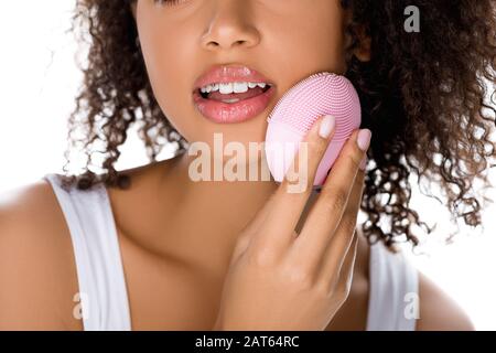 vista ritagliata della ragazza afroamericana con occhi chiusi utilizzando la spazzola per il viso in silicone, isolata su bianco Foto Stock