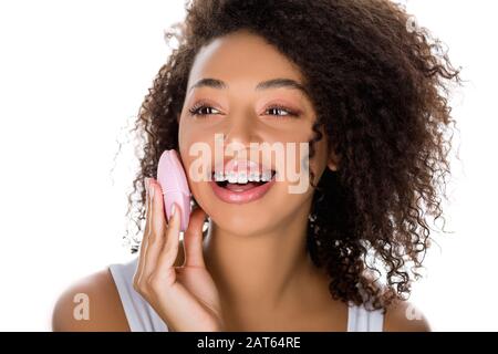 ragazza afroamericana sorridente con bretelle dentarie usando la spazzola facciale di pulizia del silicone, isolata su bianco Foto Stock