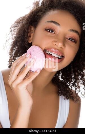 ragazza afro-americana felice con bretelle dentarie usando la spazzola facciale di pulizia del silicone, isolata su bianco Foto Stock