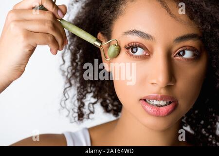 bella ragazza afroamericana curly utilizzando il rullo viso jade, isolato su grigio Foto Stock