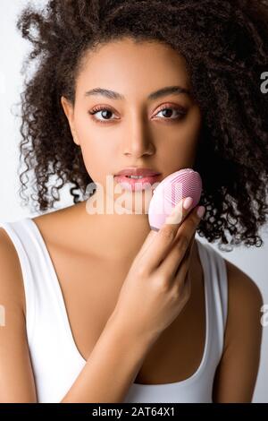 ragazza afroamericana attraente usando spazzola facciale di pulizia del silicone, isolata su grigio Foto Stock