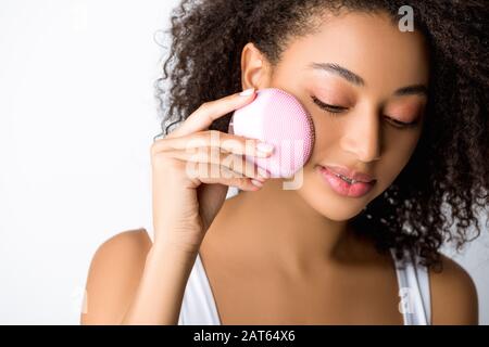 ragazza afroamericana sorridente con occhi chiusi usando spazzola facciale di pulizia del silicone, isolata su grigio Foto Stock