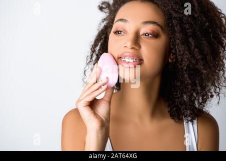 ragazza afroamericana sorridente con bretelle dentarie usando la spazzola facciale di pulizia del silicone, isolata su grigio Foto Stock