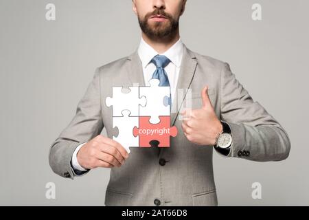 vista ritagliata di un uomo d'affari che tiene un puzzle con scritte sul copyright e che mostra il pollice isolato sul grigio Foto Stock