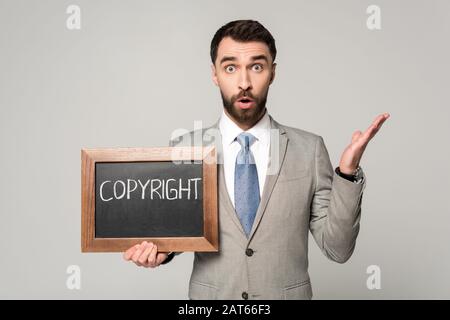 un uomo d'affari scioccato che guarda la macchina fotografica mentre tiene la lavagna con l'iscrizione di copyright isolata sul grigio Foto Stock