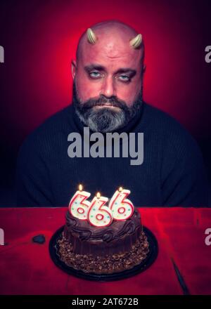 Un uomo triste siede davanti ad una torta di compleanno del cioccolato del diavolo con il numero 666 candele con uno sfondo rosso acceso Foto Stock