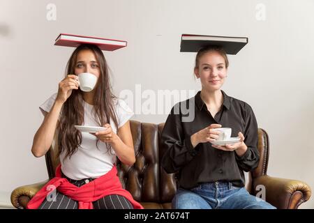 Due giovani donne che bevono caffè e libri equilibranti sulla testa. Scatto medio. Guardando la fotocamera. Foto Stock