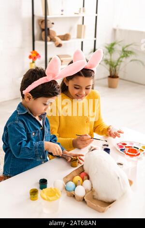 Carino fratello e sorella in orecchie conigliate che dipingano le uova di Pasqua mentre si siede alla scrivania vicino al coniglio bianco Foto Stock