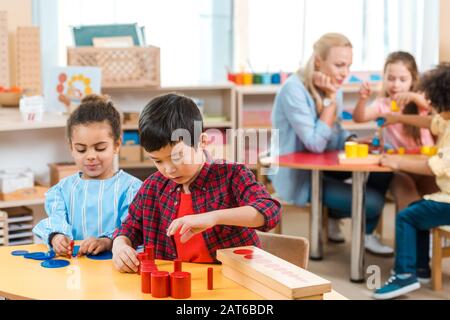 Focus selettivo dei bambini che giocano con i bambini e insegnante in background nella scuola di montessori Foto Stock