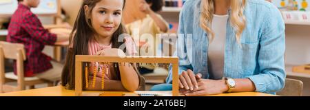 Fuoco selettivo di capretto che guarda la macchina fotografica mentre gioca il gioco educativo dall'insegnante nella scuola di montessori, colpo panoramico Foto Stock