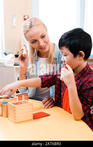 Fuoco selettivo di insegnante sorridente che guarda il capretto asiatico durante la lezione alla scuola di montessori Foto Stock