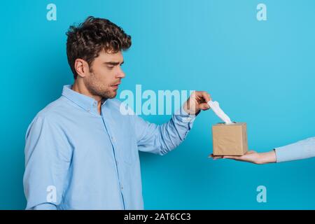 vista parziale della donna che dà pacchetto di tovaglioli di carta a uomo triste su sfondo blu Foto Stock