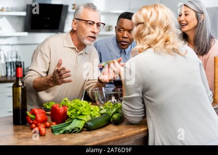 amici multiculturali sorridenti che parlano e stanno accanto al tavolo in cucina Foto Stock