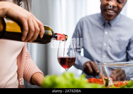 vista corta della donna che versa il vino al bicchiere e dell'uomo afroamericano che lo guarda Foto Stock