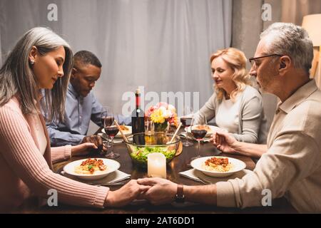 amici multiculturali che tengono le mani e pregano durante la cena Foto Stock