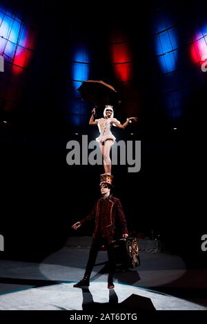 attraente ginnastica con ombrello che si esibisce mentre si alza sul cappello dell'uomo in circo Foto Stock