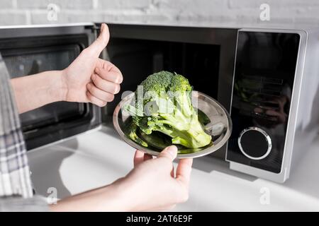 vista corta della piastra di tenuta della donna con broccoli vicino al forno a microonde e mostra come Foto Stock