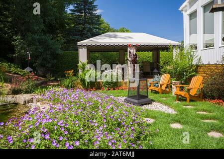 Sentiero in pietra grigia accanto alle sedie in legno Adirondack e gazebo con sedie a sdraio in vimini e laghetto delimitato da geranio, fiori 'Johnson's Blue' Foto Stock