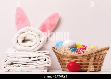 Un primo piano di una pila di lenzuola bianche pulite, asciugamani e orecchie di coniglio di Pasqua, un cesto di uova su un tavolo, su uno sfondo di pareti chiare. Foto Stock