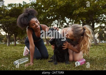 Due giovani e sorridenti amici atletici donne che giocano e amano un cane nero nel parco Foto Stock