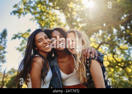 Tre giovani donne sorridenti multietniche in abbigliamento casual stare insieme sotto la luce del sole al parco avendo divertimento e ridere Foto Stock