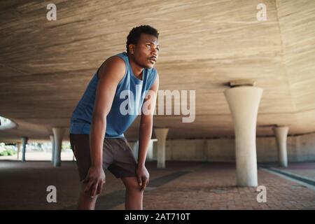 Corridore maschio esausto che prende un riposo dopo aver eseguito duro sulla strada sotto un ponte di cemento Foto Stock