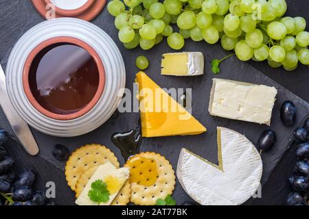 Tagliere di formaggio con formaggio assortito, camembert, cheddar, gouda, cracker, uva e vaso di miele su sfondo nero ardesia - foto in alto Foto Stock