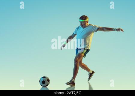 Uomo anziano che gioca a calcio, calcio su sfondo gradiente in luce al neon. Il modello maschio caucasico in grande forma rimane attivo, sporitivo. Concetto di sport, attività, movimento, benessere, stile di vita sano. Foto Stock