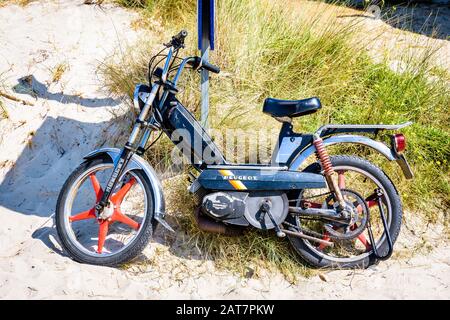 Un ciclomotore nero Peugeot 103 SP d'epoca della 1980s con ruote rosse Rigida Leleu a 5 razze appoggiate contro una pole al sole in spiaggia. Foto Stock