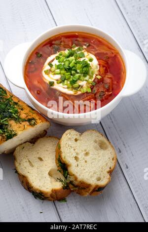 Borsct rosso russo con panna acida e verdure in ciotola bianca e pezzi di pane su sfondo di legno, tavolo. Concetto di cucina Ucraina Foto Stock