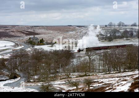 Treno d'epoca che emette fumo che taglia attraverso le North York Moors fiancheggiata da alberi in un paesaggio innevato in inverno vicino Goathland, Yorkshire, UK. Foto Stock