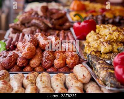 Varietà di carne alla griglia al negozio di cibo di strada, maiale, manzo, pile di salsicce di pollo e peperoni rossi su vassoi. Malsano oleoso barbeque strada fast food per Foto Stock